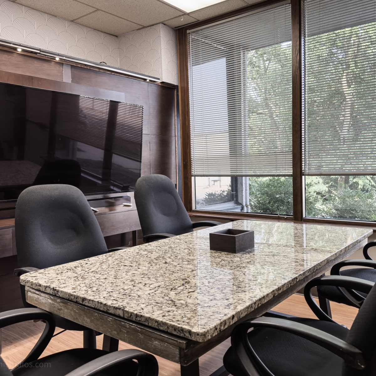 A conference room with a granite table surrounded by black chairs, featuring a television on the wall and windows with blinds.