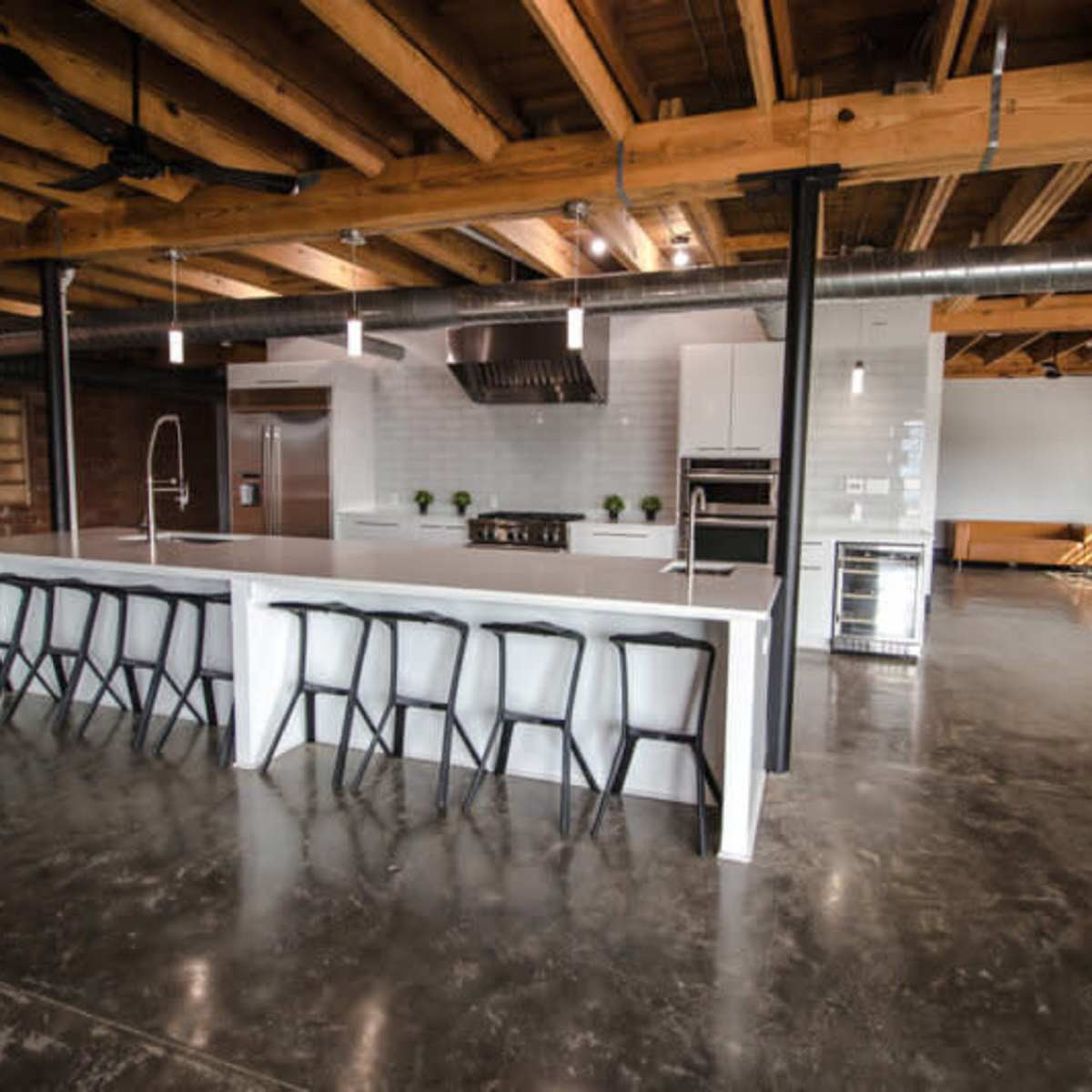 A spacious modern kitchen with a large white island, eight black bar stools, stainless steel appliances, and exposed wooden beams.