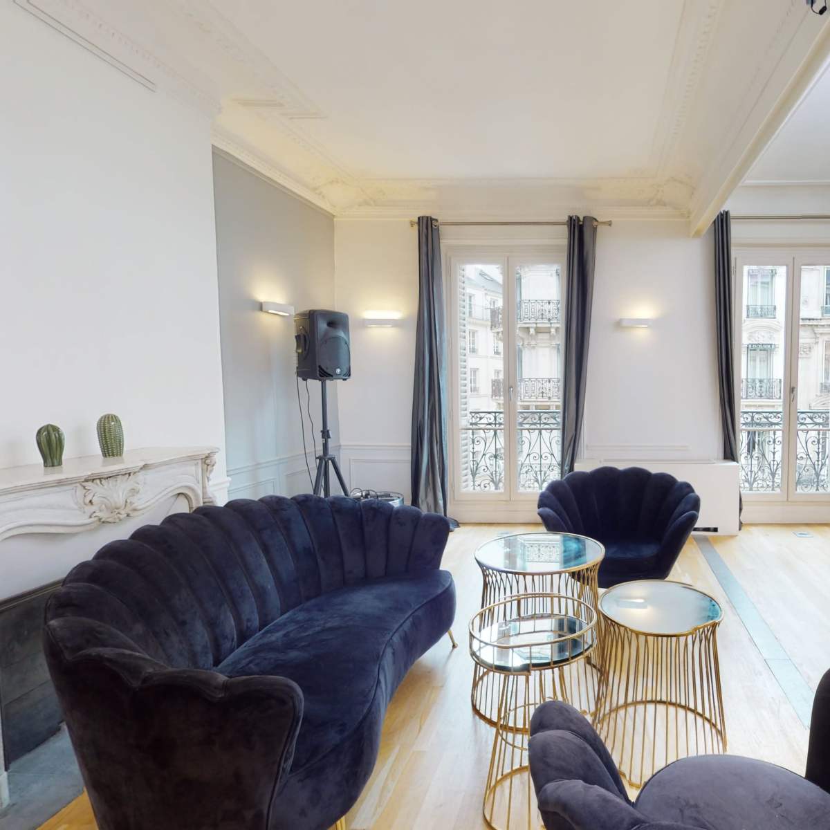 A spacious room featuring a dark velvet sofa, gold and glass side tables, and large windows with draped curtains.