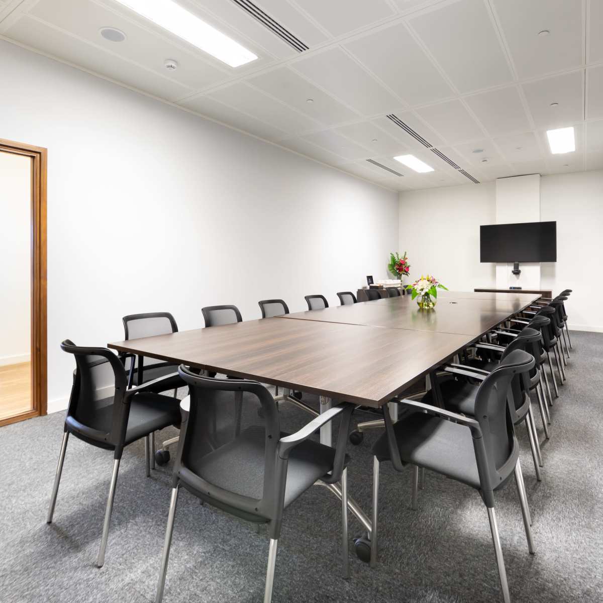 A large conference room features a long wooden table surrounded by black chairs, with a television on one wall and an adjoining glass door.