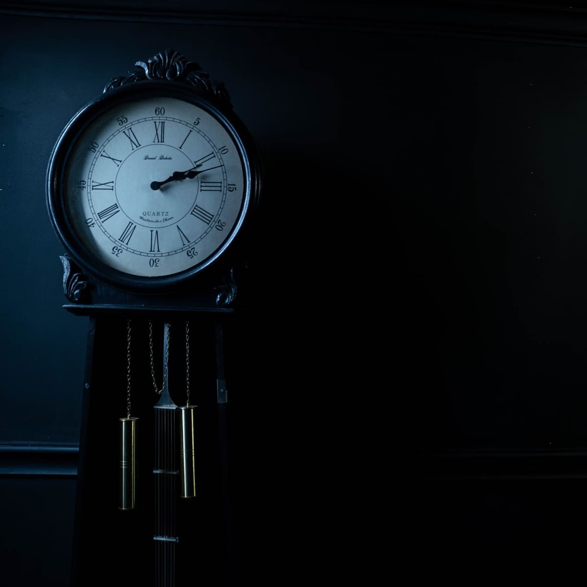 A grandfather clock with a circular face and pendulum stands against a dark wall.