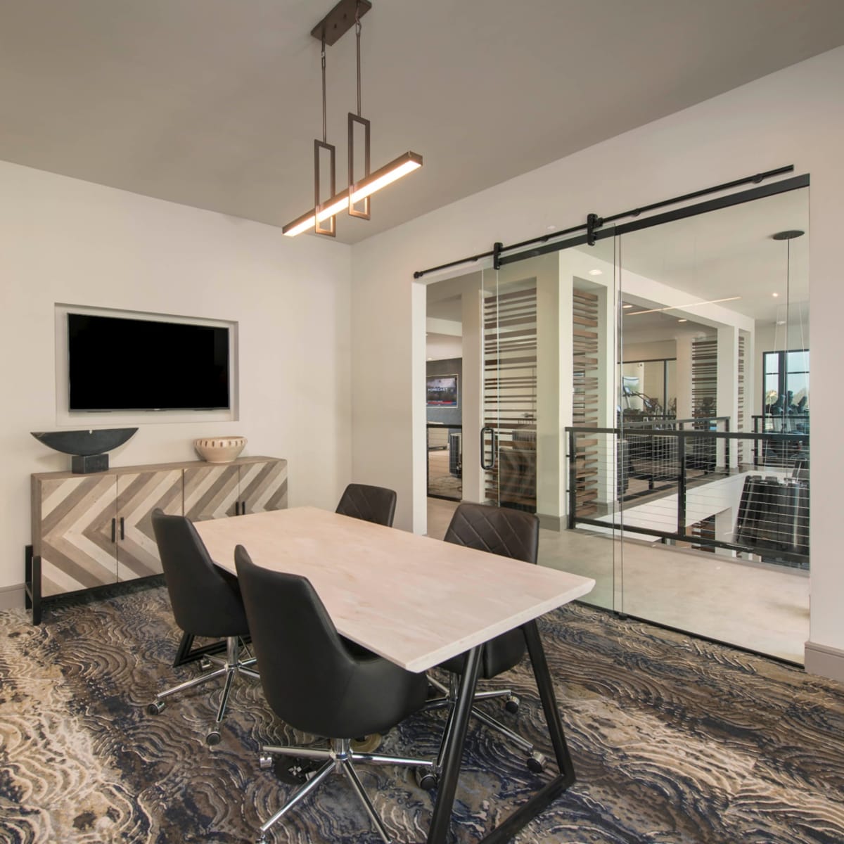 A modern conference room features a rectangular table surrounded by black chairs, with a television mounted on the wall and large glass doors leading to an adjacent space.