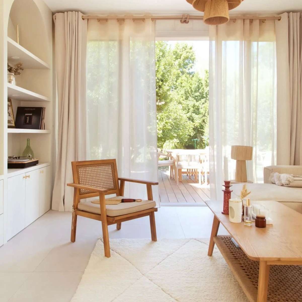 A cozy living room features a wooden armchair, a light wooden coffee table, and large windows displaying an outdoor deck surrounded by greenery.