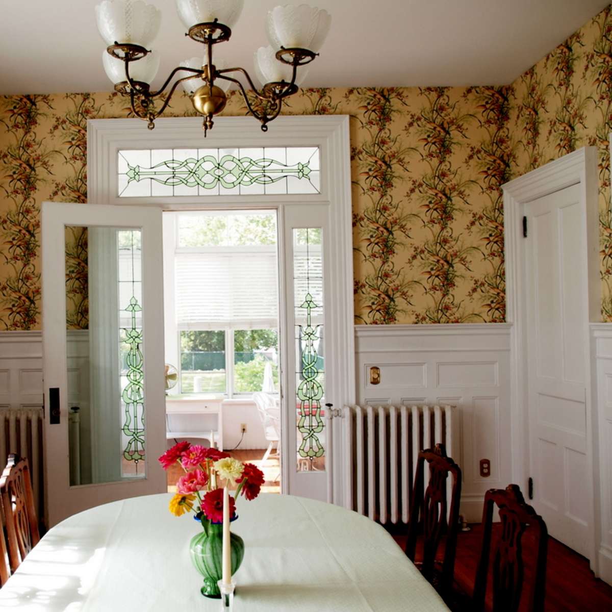 A dining room with a floral wallpaper, a large table at the center, a vase of flowers, and a door leading to a sunlit area.