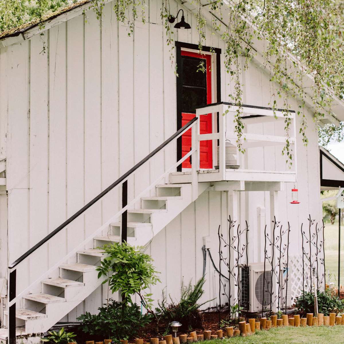 A white wooden house features a red door and a staircase leading to it, surrounded by a garden with potted plants.