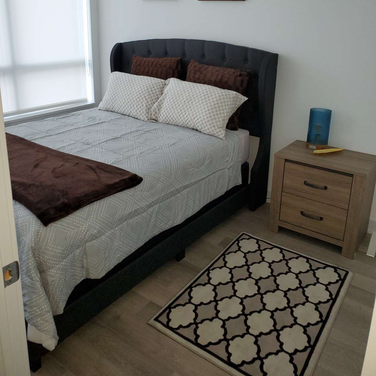 A neatly made bed with decorative pillows in a well-lit bedroom, accompanied by a nightstand and framed artwork on the walls.