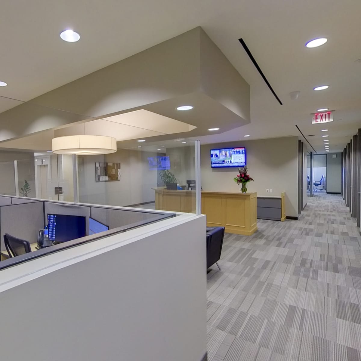 A modern office interior featuring cubicles, a reception area with a desk, and a seating space, all brightly lit by overhead lighting.