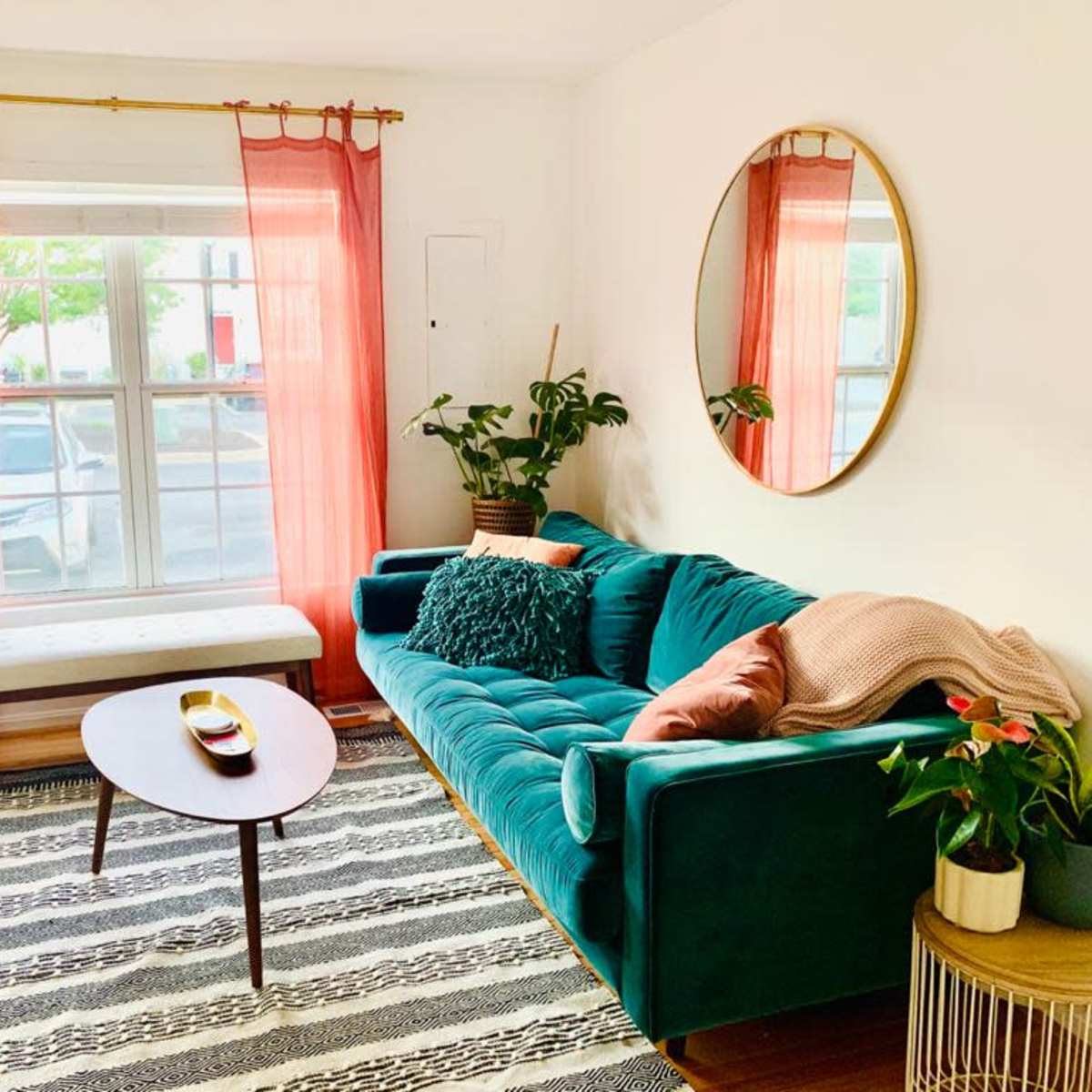 A cozy living room features a teal sofa, a round mirror, pink curtains, and a coffee table surrounded by plants and chairs.