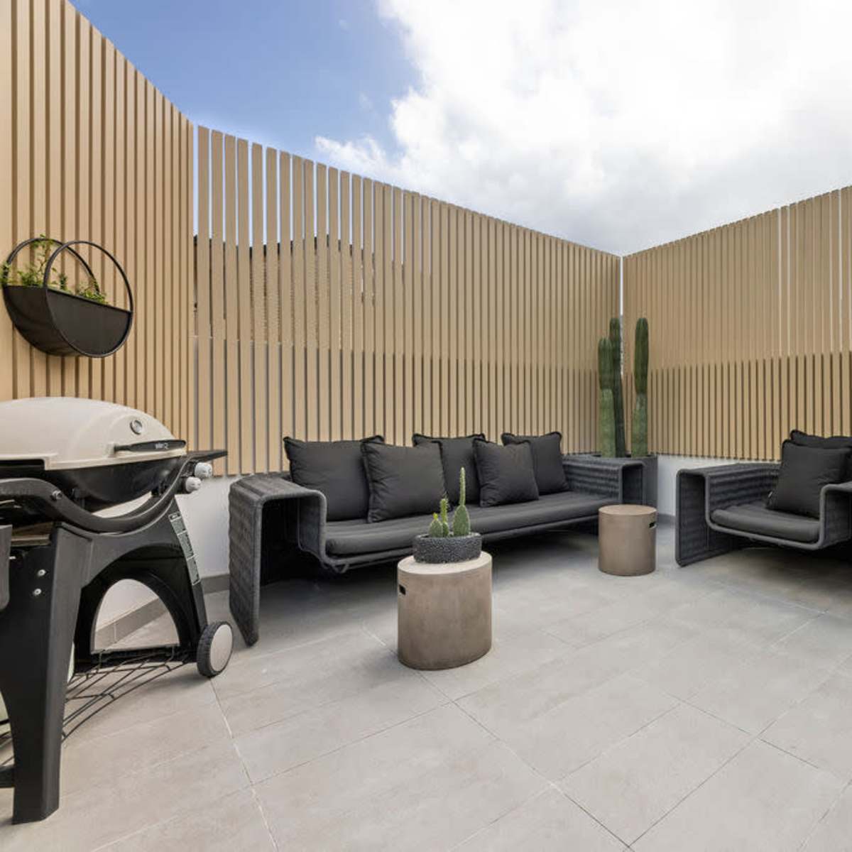 A modern outdoor terrace featuring a black sectional sofa, a small round table, a barbecue grill, and potted plants, all surrounded by wooden slat fencing.
