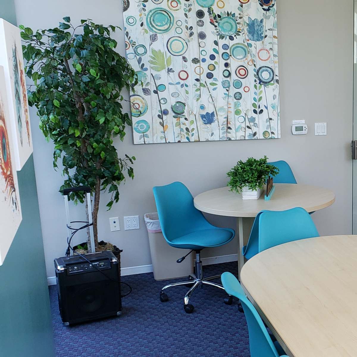 A modern office space featuring two round tables with teal chairs, decorative wall art, and a plant in a corner.