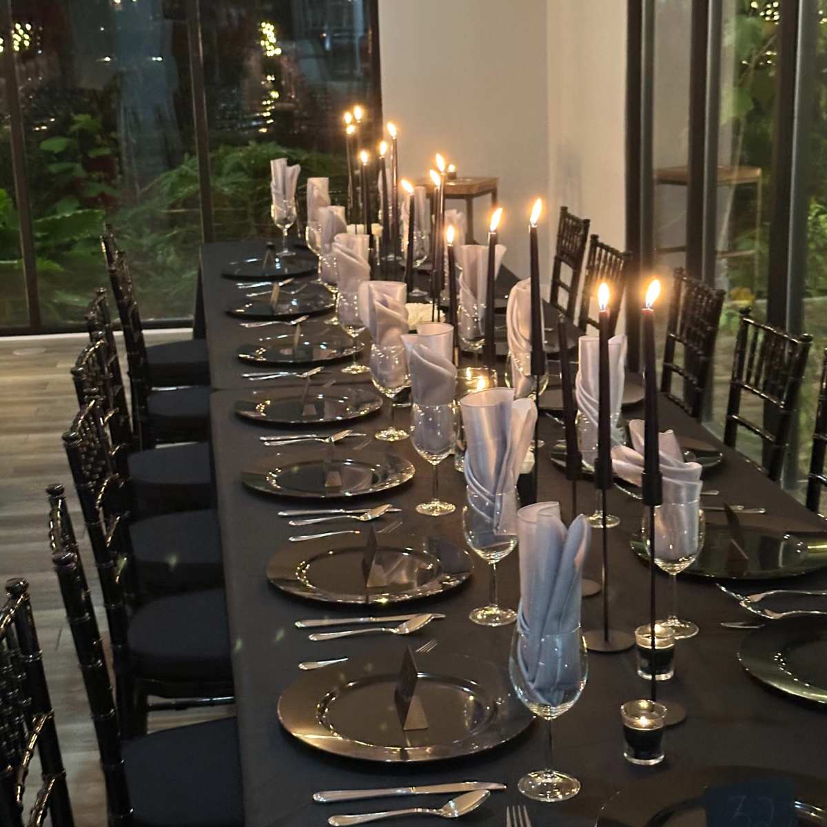 A long table is set for a formal dinner, featuring black tablecloths, lit candles in holders, and neatly arranged plates and utensils.