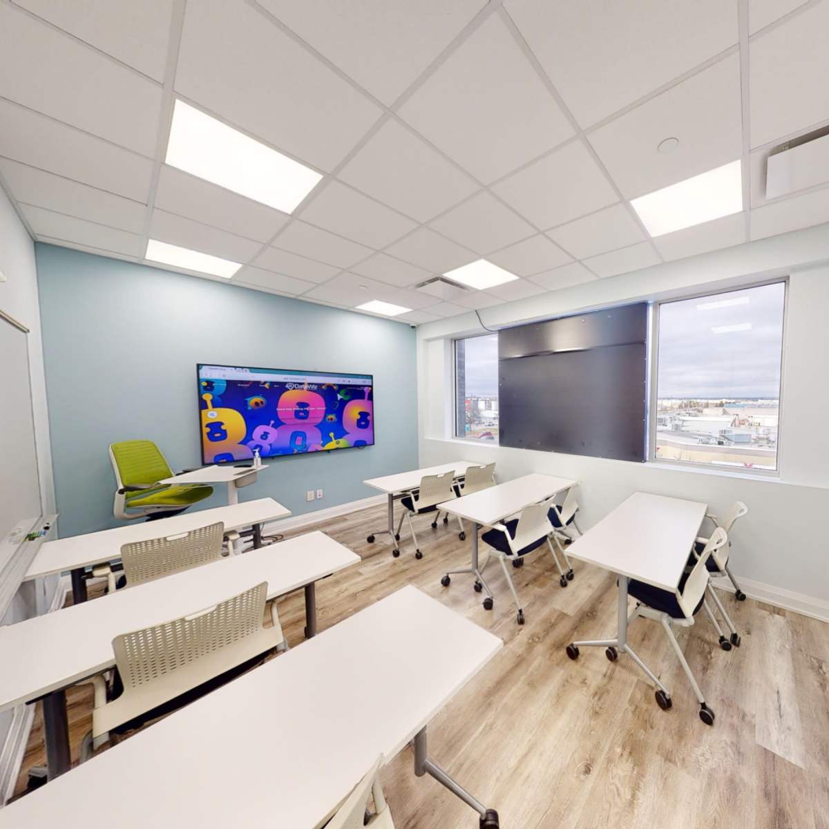 A modern classroom features several movable desks and a large screen on the wall, with large windows allowing natural light to enter.