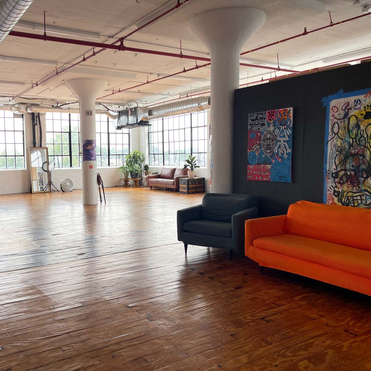 A spacious, industrial-style room with wooden floors, large windows, and colorful furniture, including a green and an orange sofa, alongside artwork on the walls.
