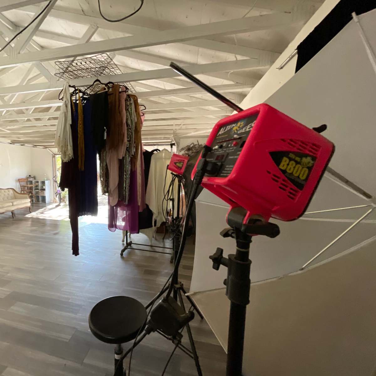 An open studio space with hanging clothing, a light stand, and a vintage couch against a white wall.