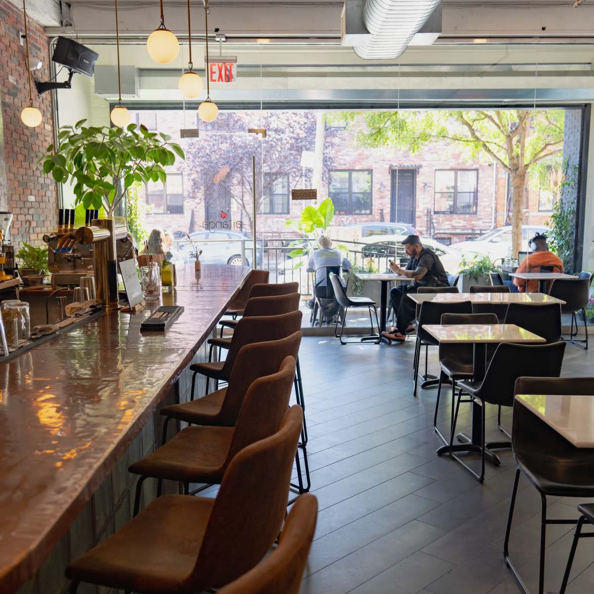 A modern café interior with a long bar on one side, featuring seating for patrons, and tables scattered throughout, with large windows overlooking a tree-lined street.