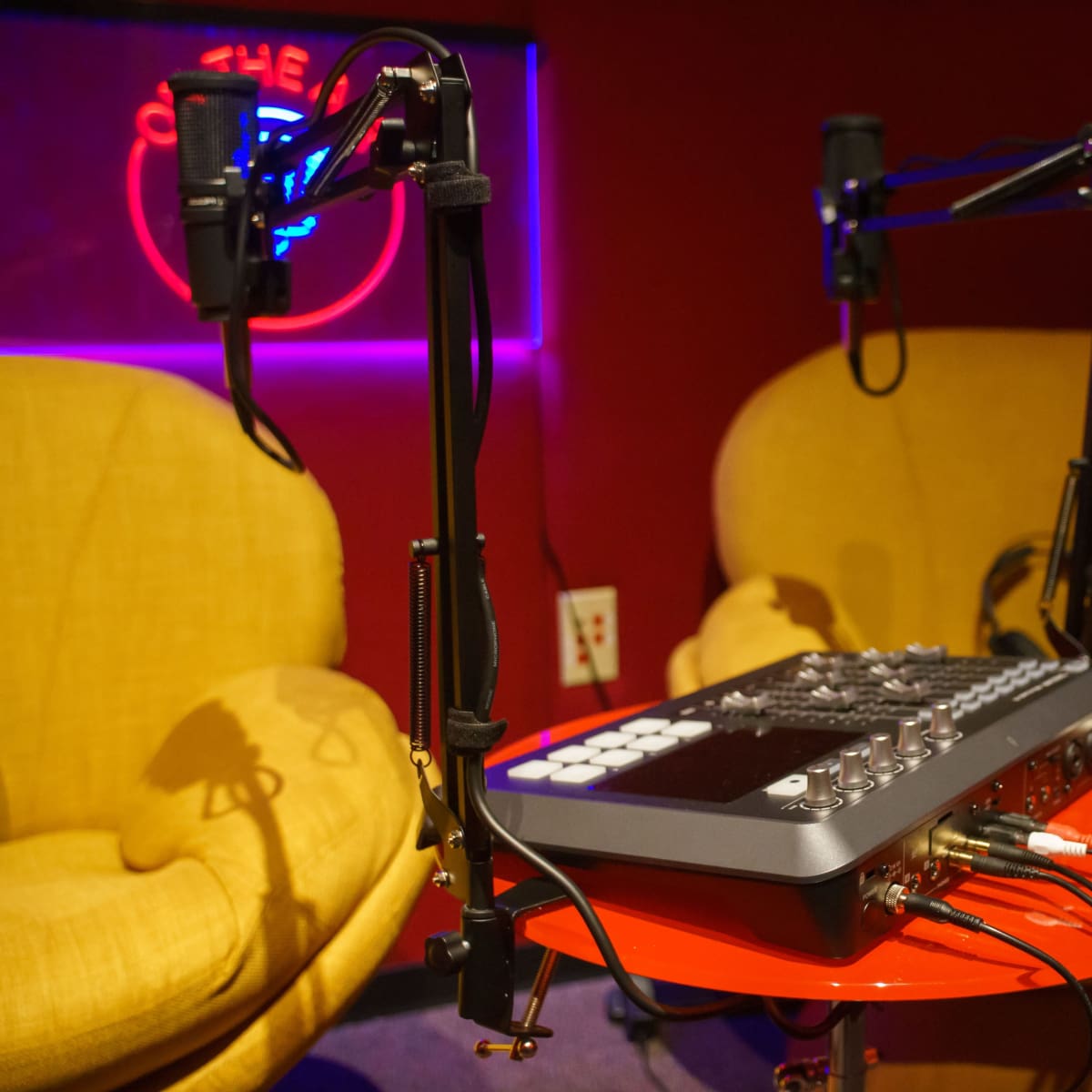 A podcasting setup featuring a mixing console with microphones on adjustable stands beside two yellow chairs against a red wall with neon lighting.