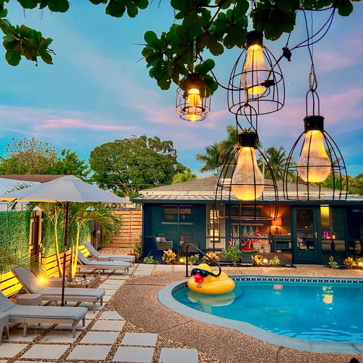 A well-lit backyard features a swimming pool surrounded by lounge chairs, a floating inflatable duck, and pendant lights hanging from overhead branches.