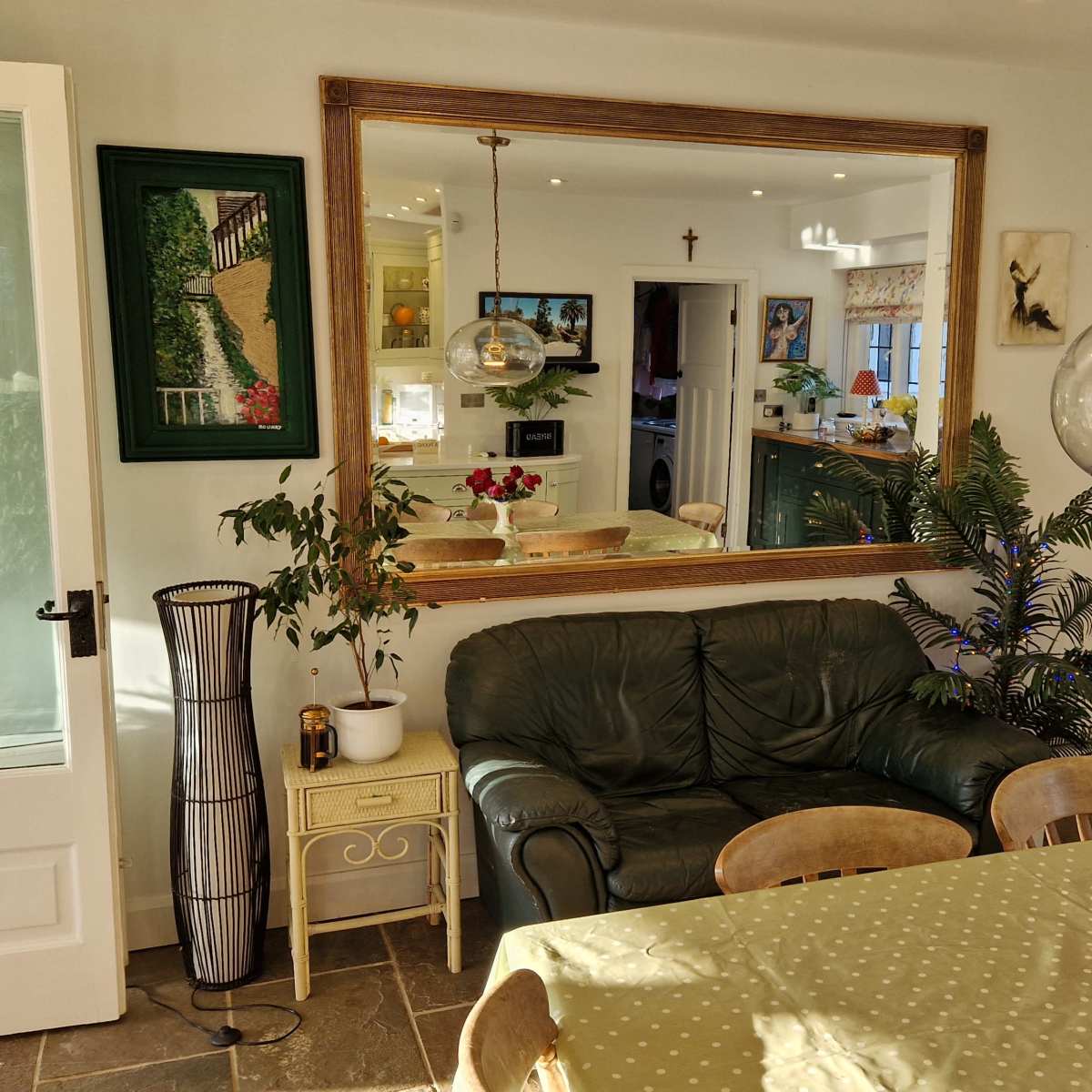 A spacious dining area features a large mirror, a green sofa, a wooden table with a polka dot tablecloth, and a vase of flowers, surrounded by various decorative elements.