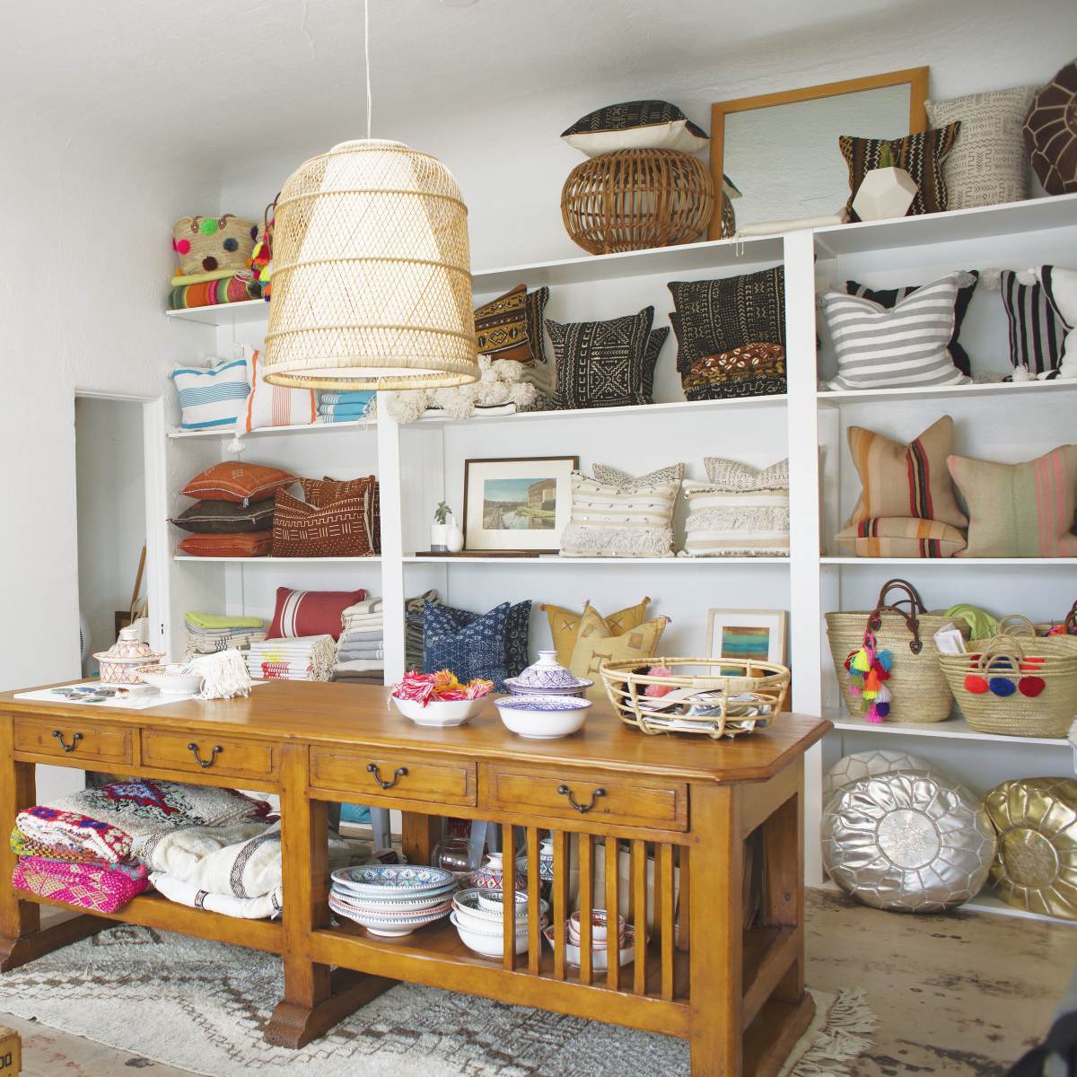A bright, organized shop interior featuring a wooden table surrounded by shelves filled with decorative pillows, woven baskets, and colorful textiles.