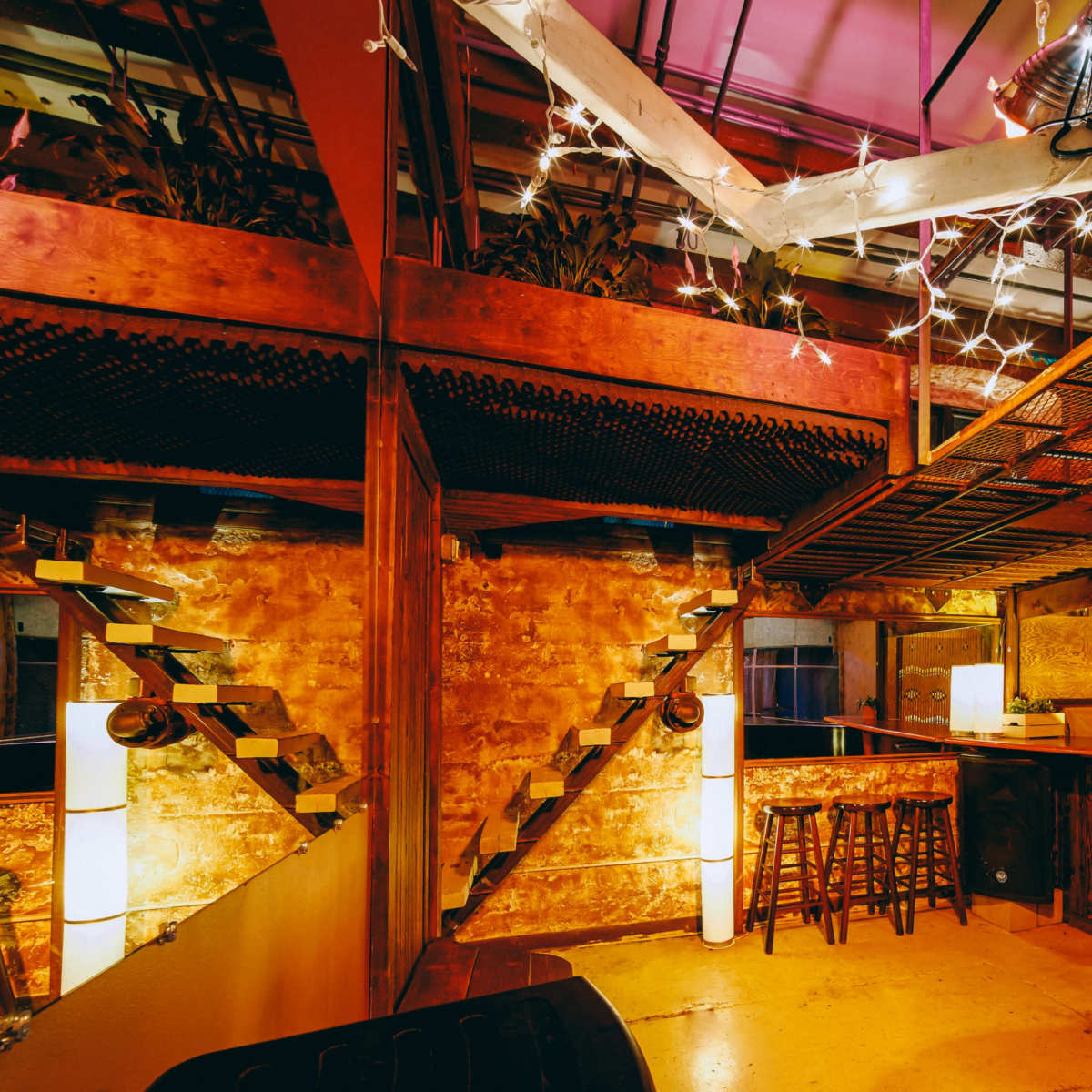 An interior space featuring a rustic bar area with stools, a piano, and a multi-level design supported by metal stairs and bright string lights.
