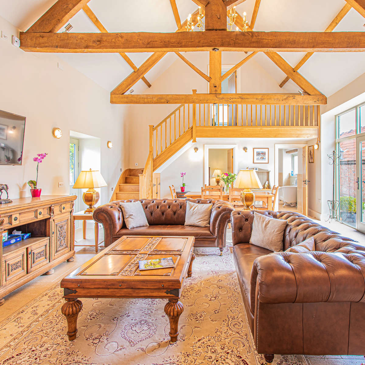 A spacious living room with a high wooden beam ceiling, a leather sectional sofa, a wooden coffee table, a large TV mounted on the wall, and natural light coming through large windows.