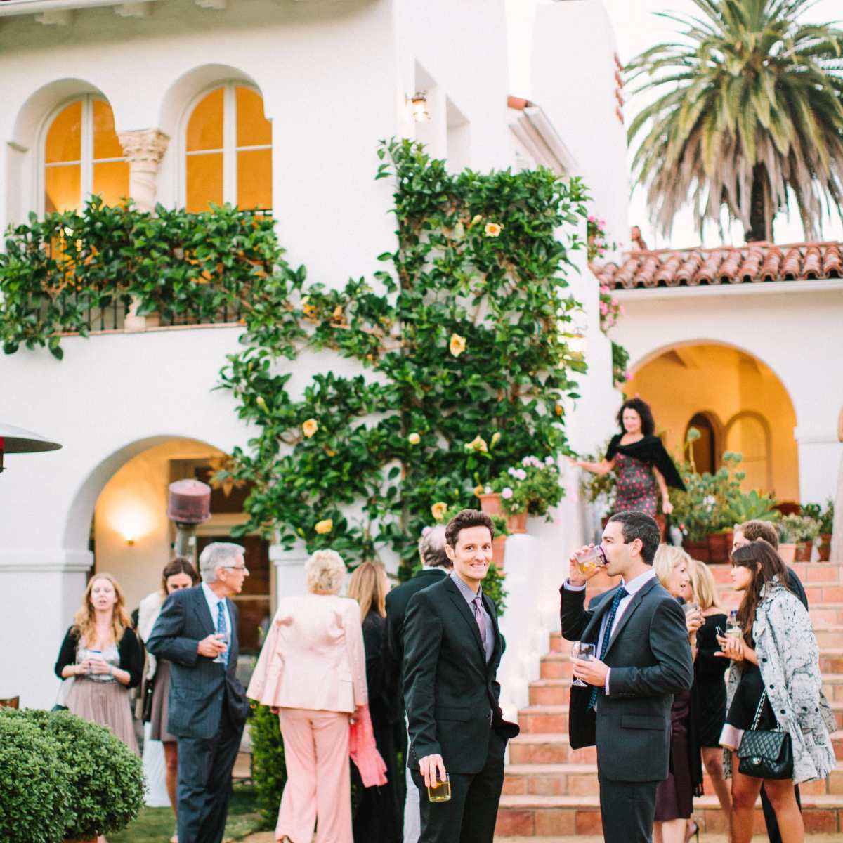 A group of people socialize on an outdoor patio in front of a stylish white building adorned with plants and palm trees.