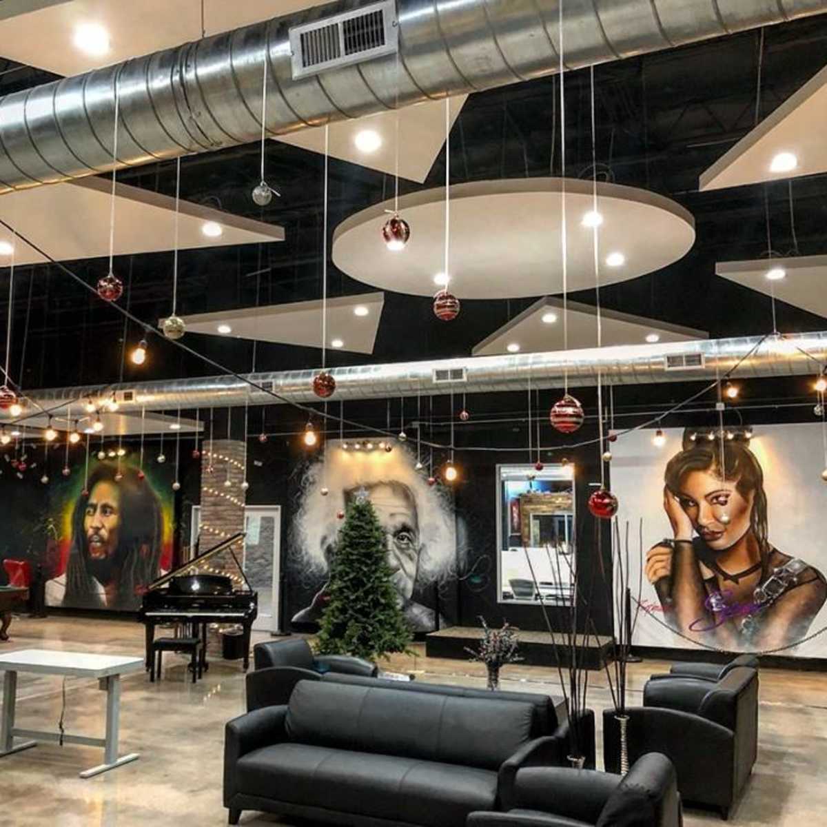 The interior of a modern studio space features a black sofa arrangement, a grand piano, and vibrant wall murals of famous figures, along with decorative lighting and a Christmas tree.