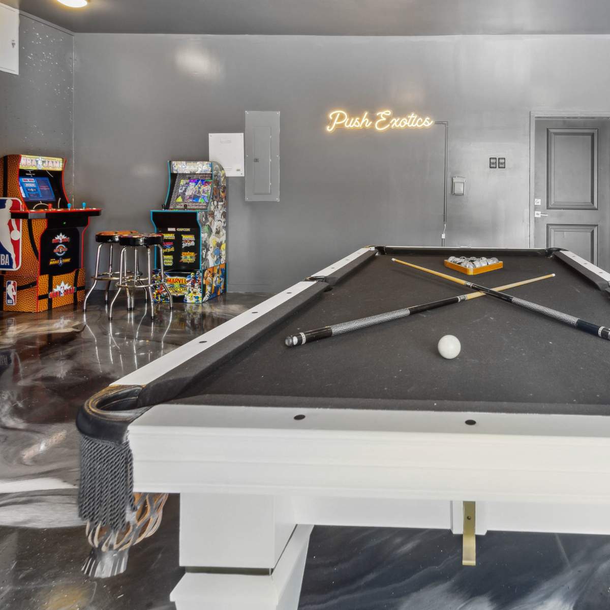 A billiard table with two cues and a single white ball is positioned in a game room featuring neon signage and retro arcade machines.