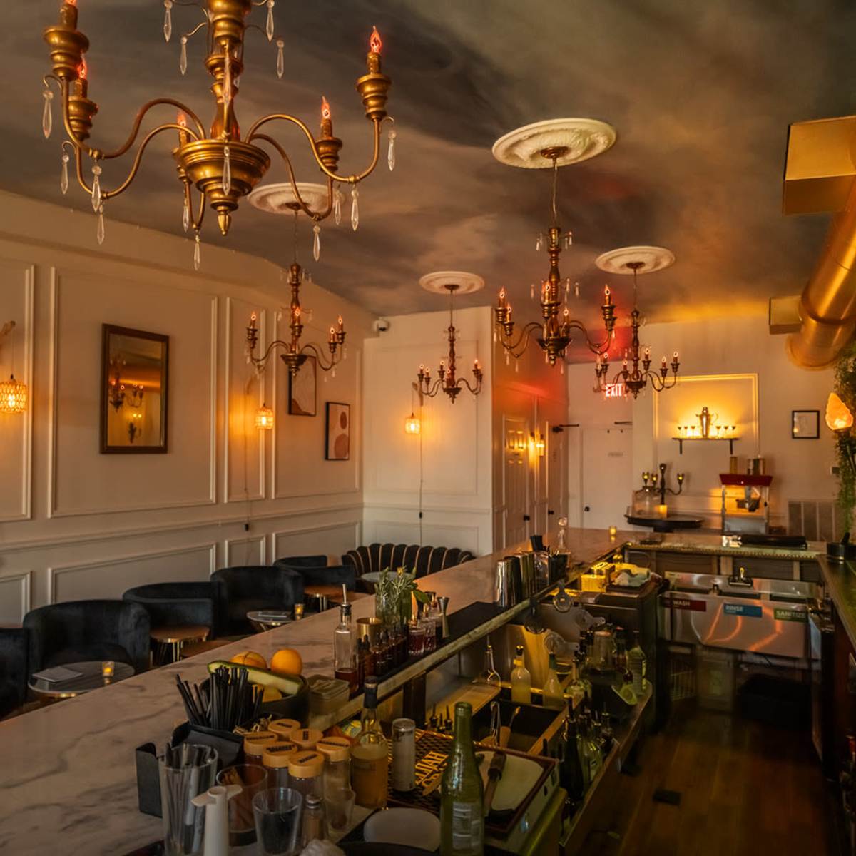 The interior of a dimly lit bar features a marble countertop, elegant chandeliers, and a well-stocked back shelf with various bottles.