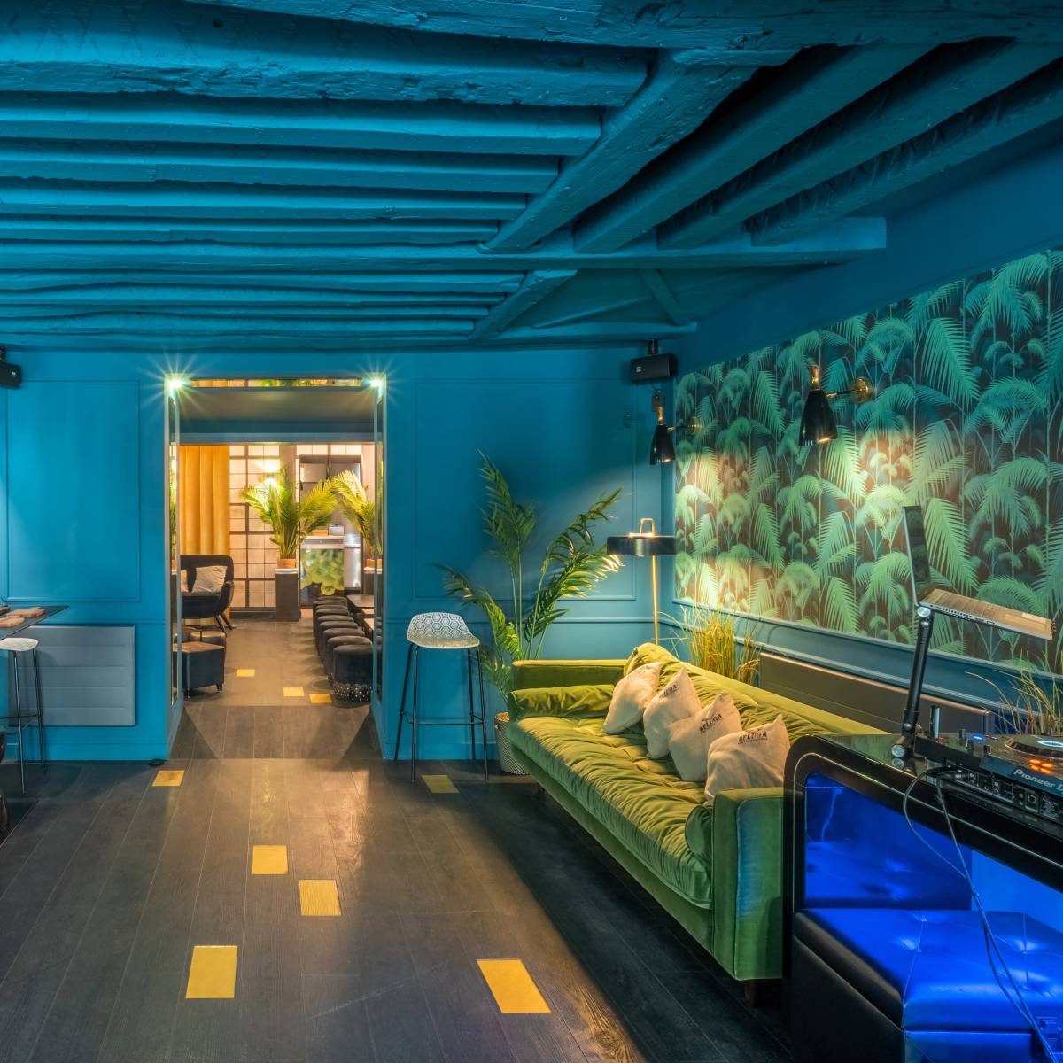 A brightly colored room with blue walls, a green sofa, and a mix of modern furniture, illuminated by soft lighting and featuring plants in the background.