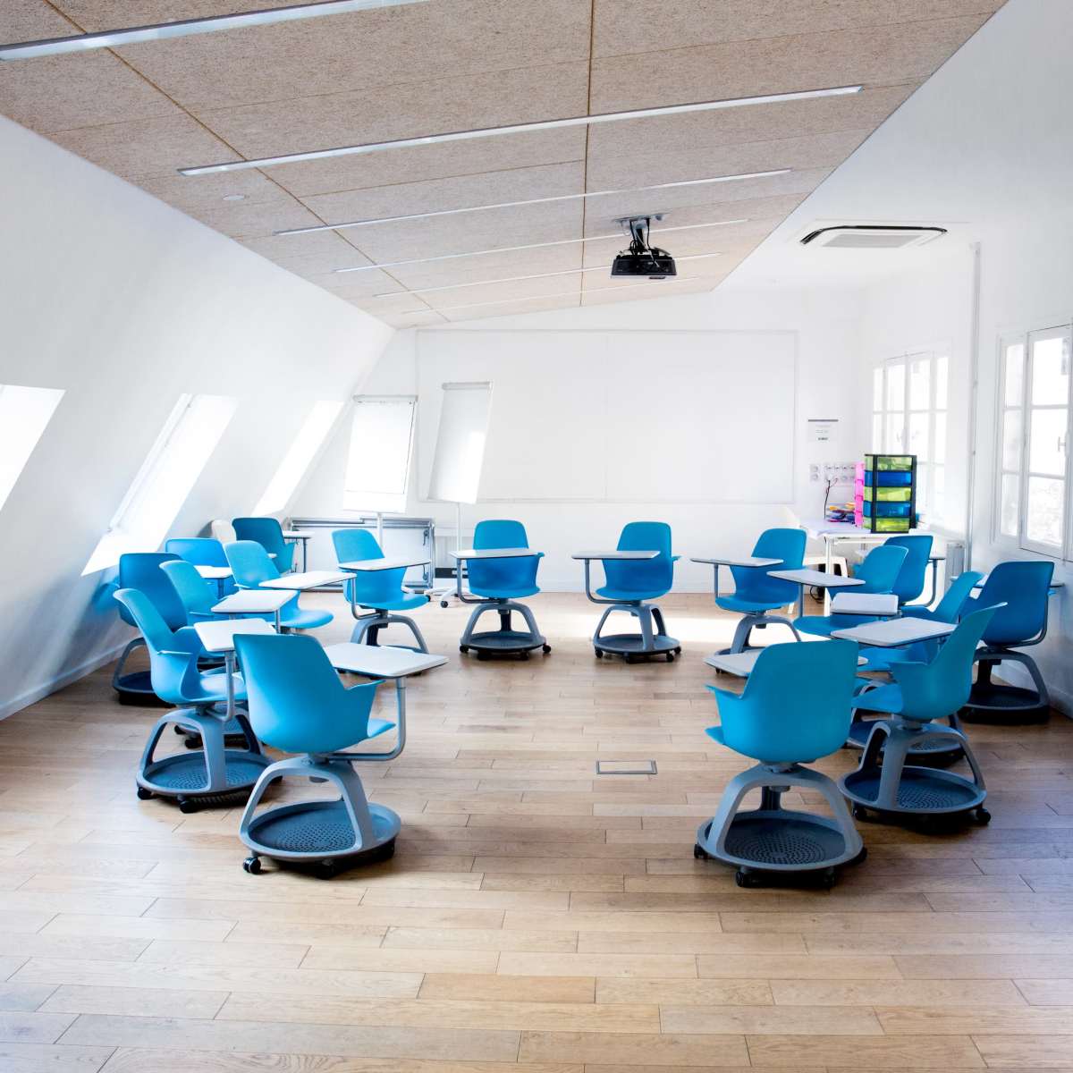 A modern classroom features circular tables and blue rolling chairs arranged around the room with large windows and a ceiling-mounted projector.