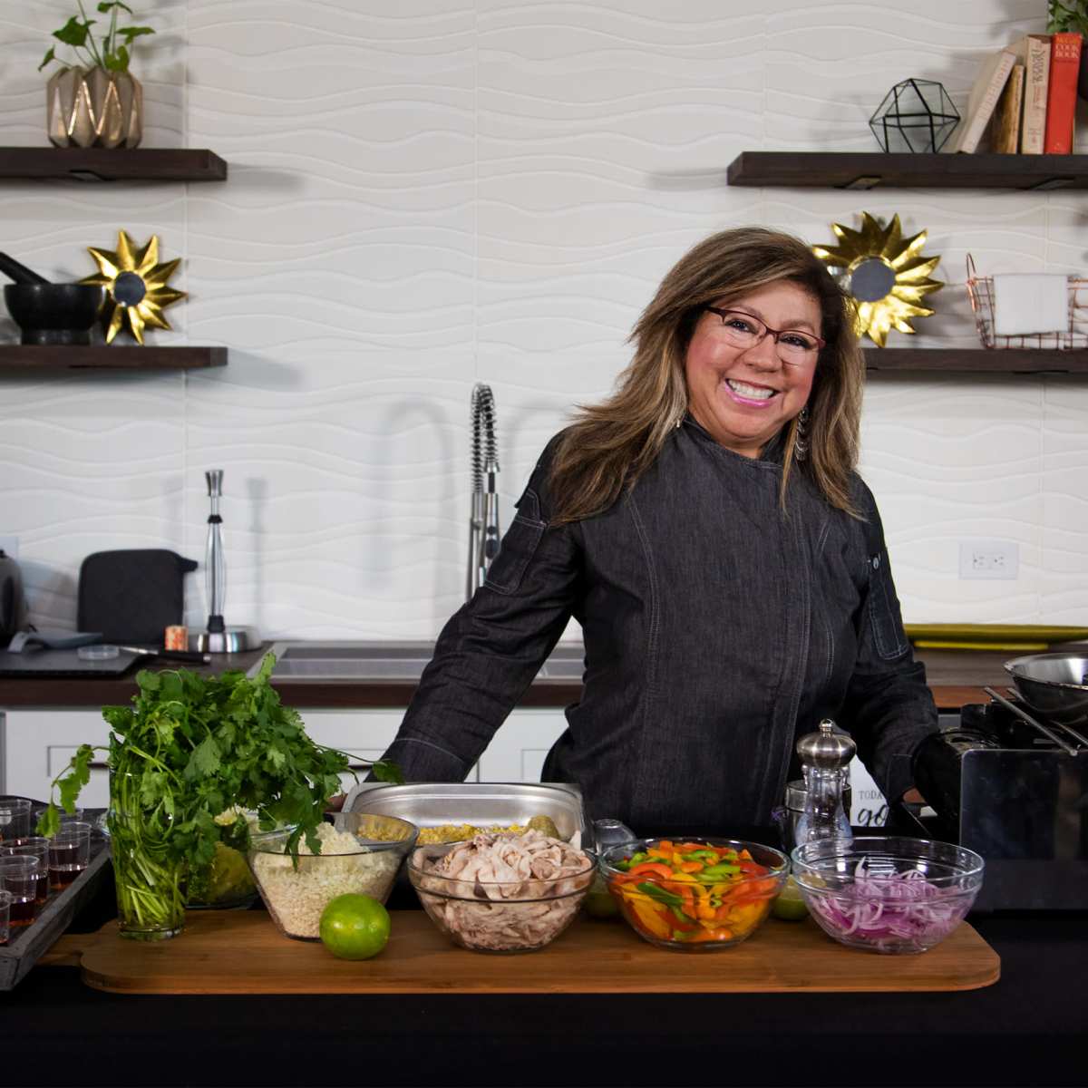 A woman stands behind a wooden table featuring various ingredients and cooking equipment in a modern kitchen setting.