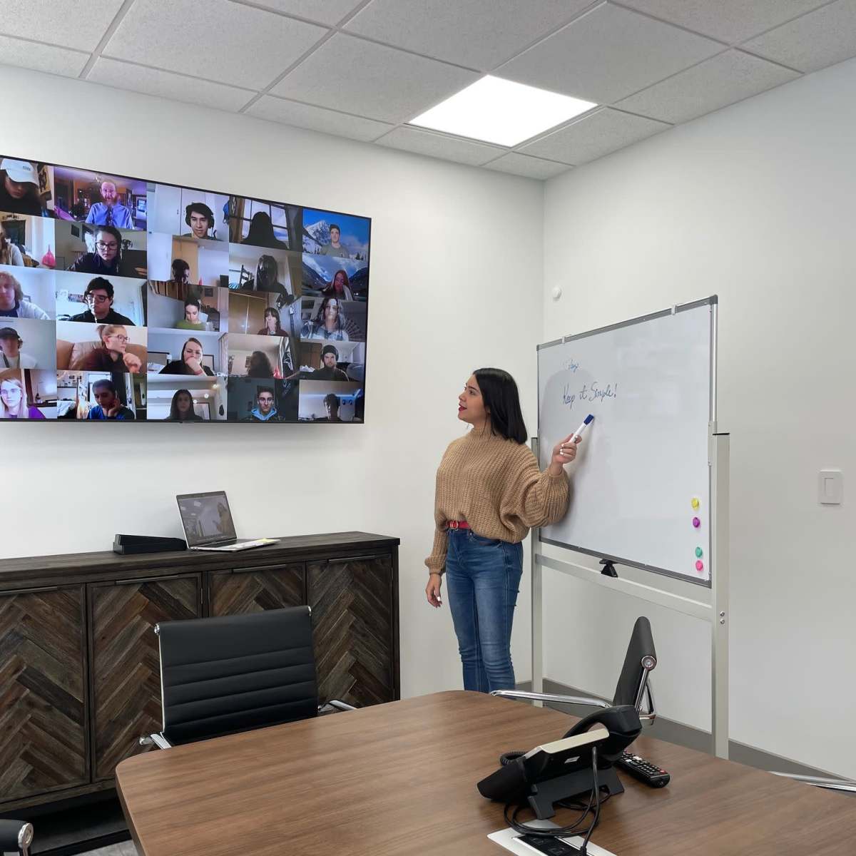 A person stands by a whiteboard in a conference room while a video call with multiple participants is displayed on a screen behind them.