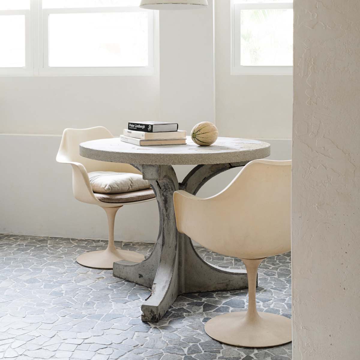 A round table with two chairs is positioned near a window, illuminated by a hanging light fixture, with books on the table and a textured floor beneath.