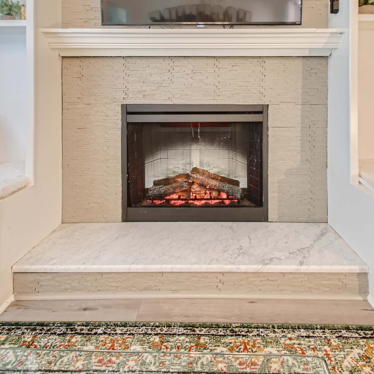 A modern fireplace with an electric insert sits beneath a wall-mounted television, framed by a light-colored stone surround and a marble base, against a backdrop of built-in shelves.