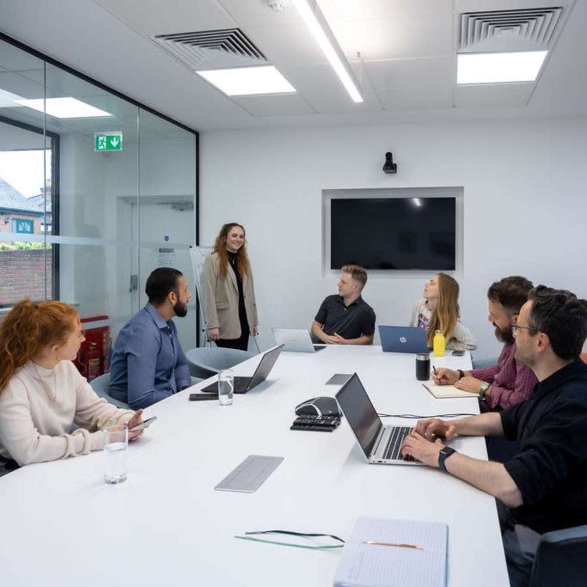A group of six people are sitting around a long, white conference table in a modern meeting room, while one person stands and addresses the group.