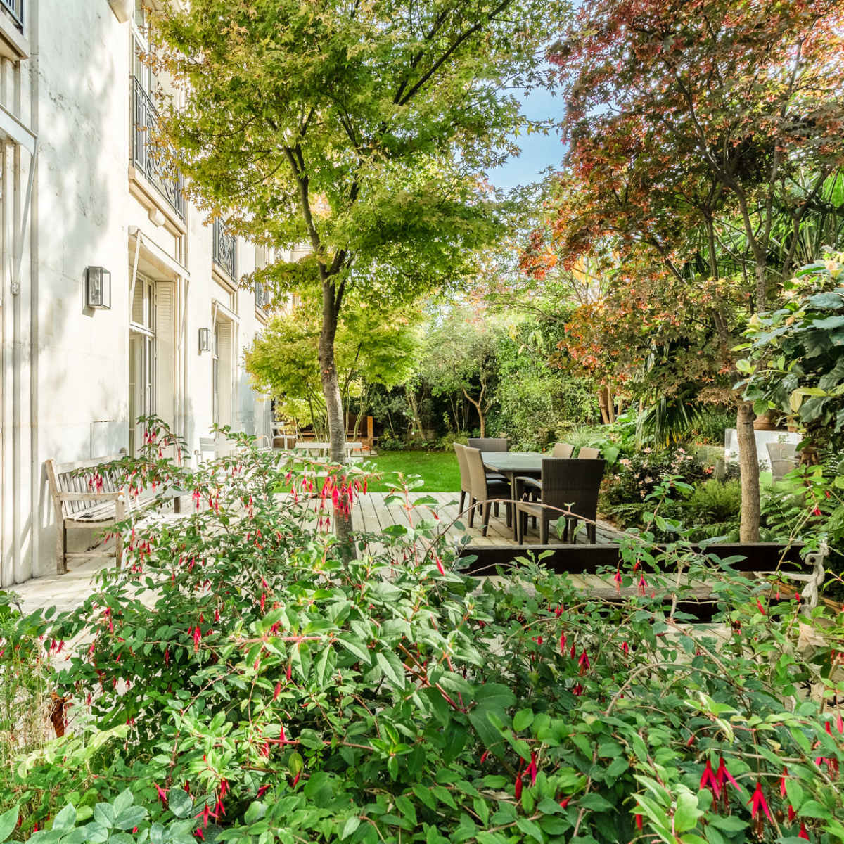 A well-maintained garden with green plants and trees beside a building, featuring outdoor seating arrangements.