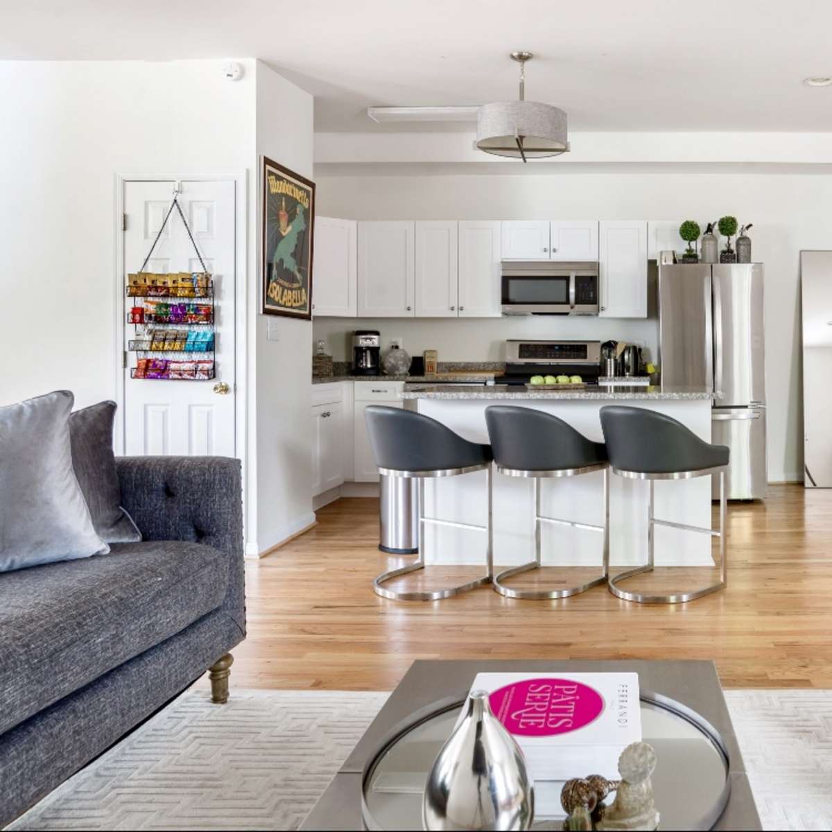 A modern living room with a gray sofa and armchair, a coffee table with a magazine, and a kitchen area featuring white cabinets and bar stools.