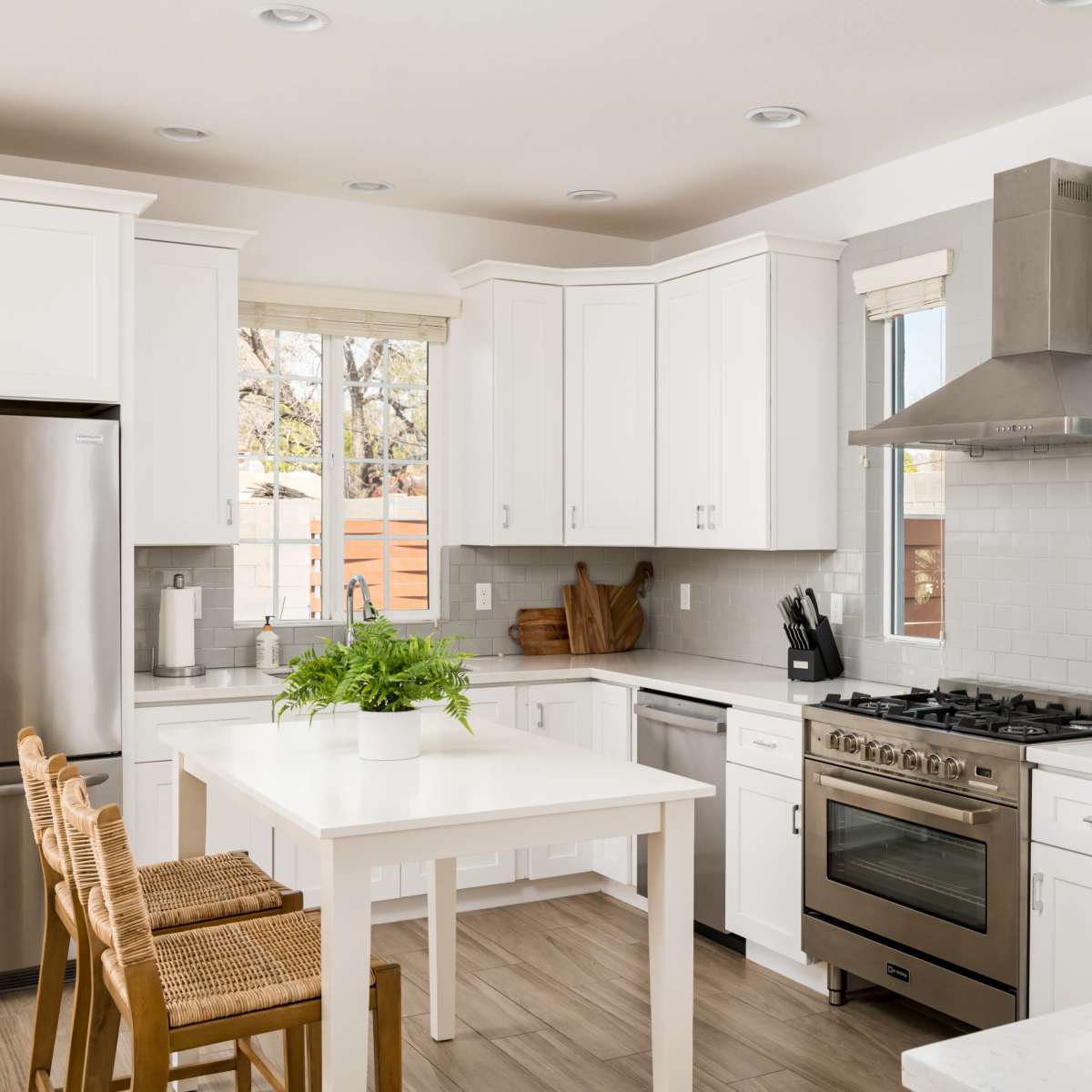 A modern kitchen features white cabinetry, stainless steel appliances, and a central table with barstools and a potted plant.