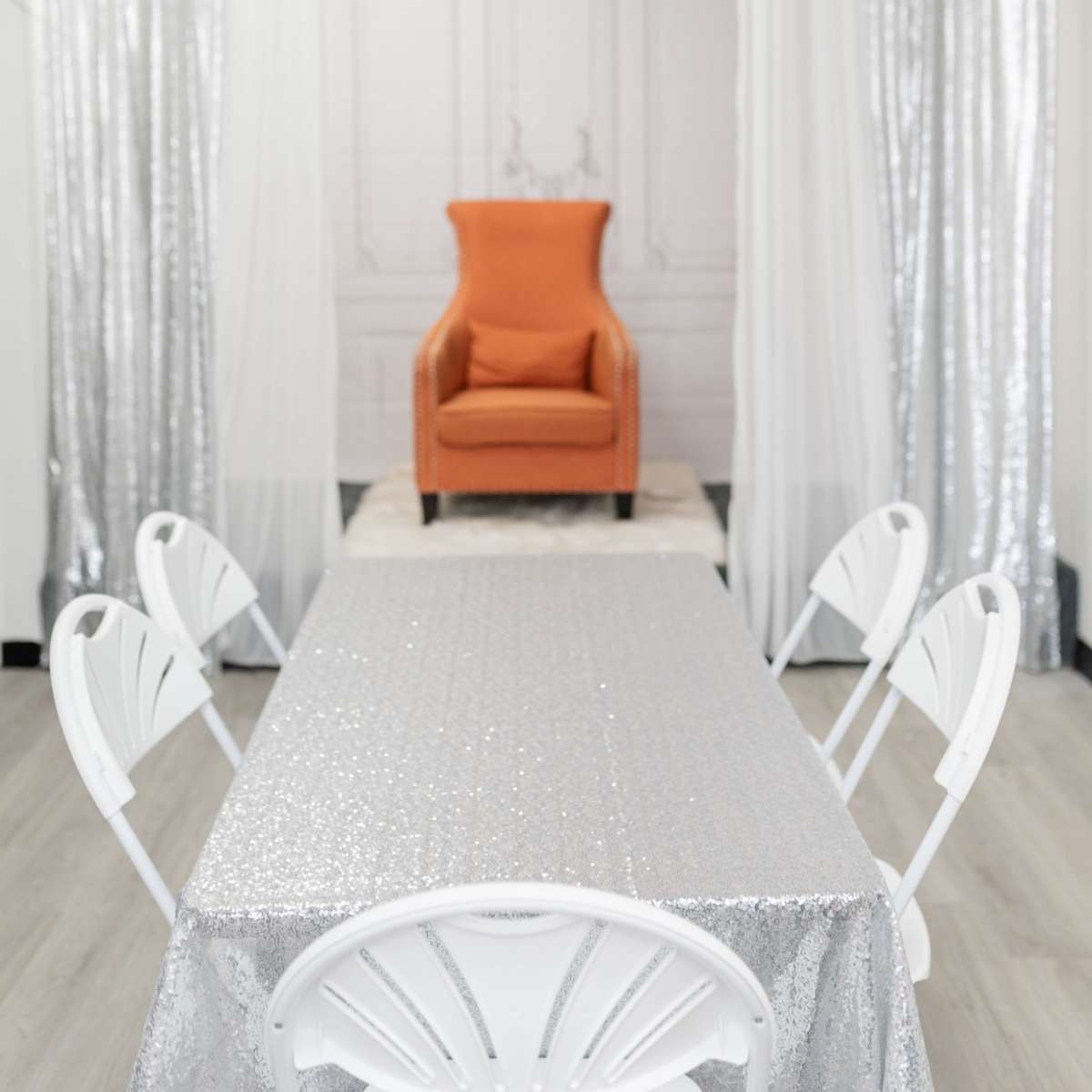 The scene features a well-lit room with a sparkling silver table and an orange armchair at the back, flanked by sheer white and silver drapes.