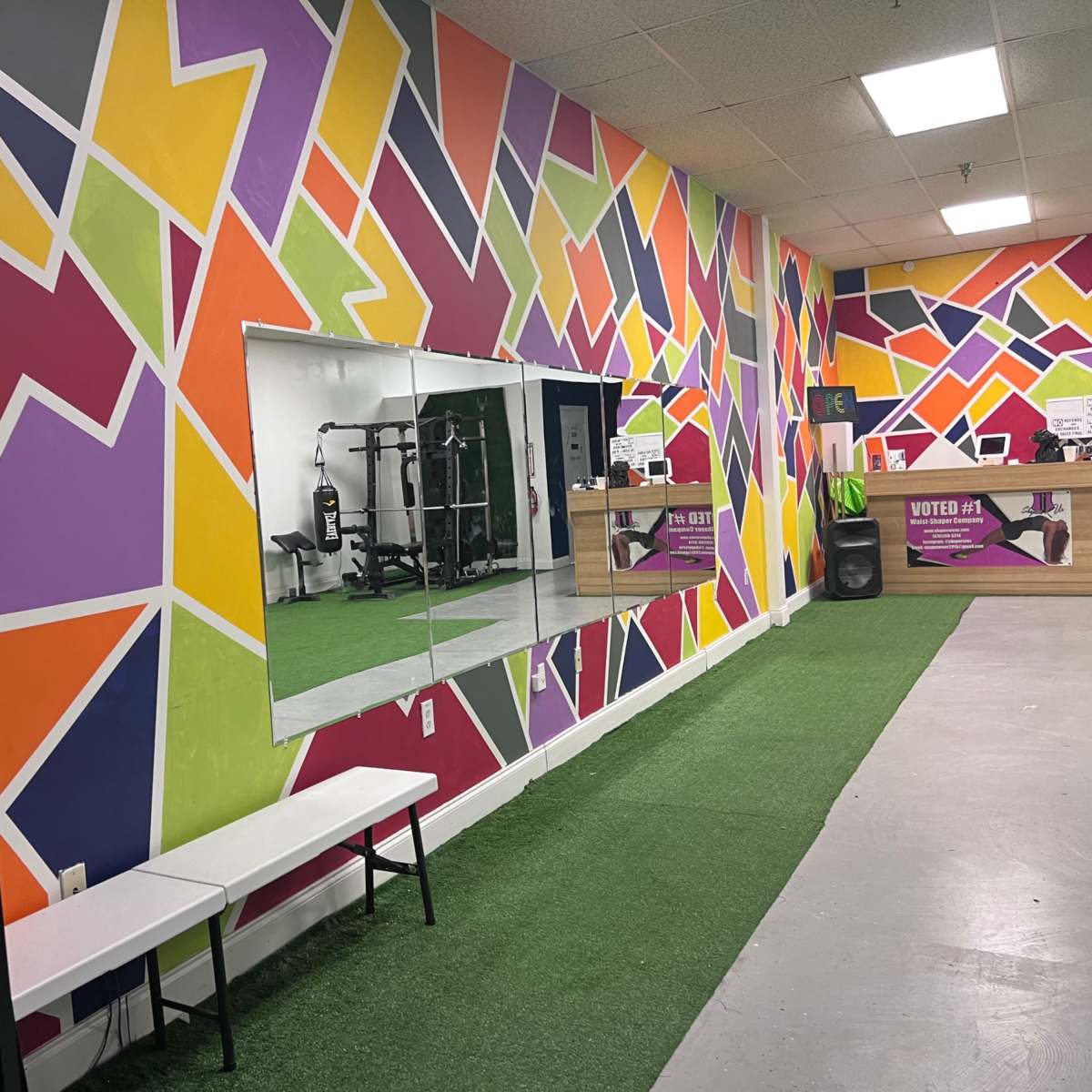 A vibrant gym interior with a colorful geometric mural on the walls, a mirror, a green turf area, and workout equipment visible in the background.