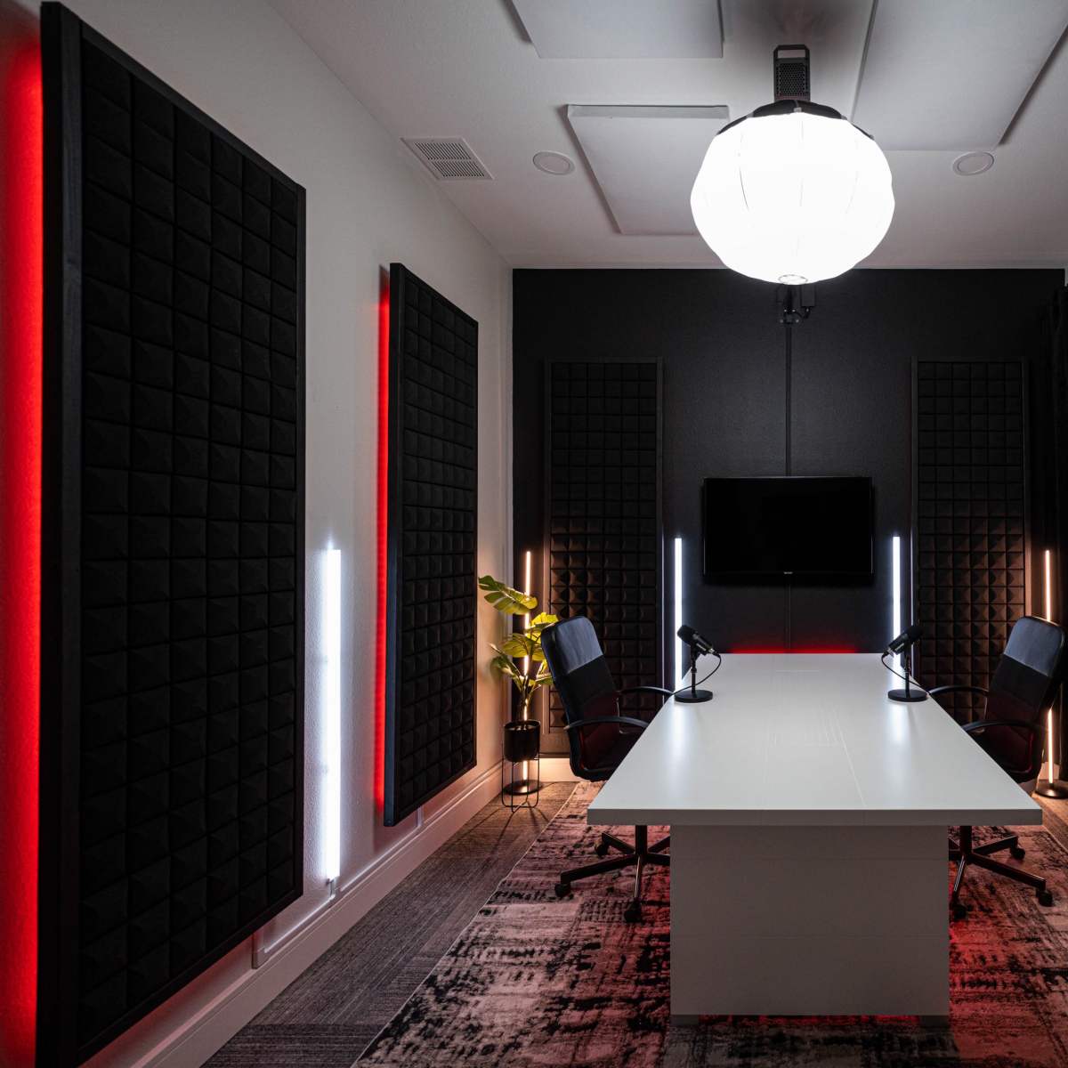 A modern conference room with two black chairs and a large white table, illuminated by a pendant light and accented with black soundproof panels and red LED lights.