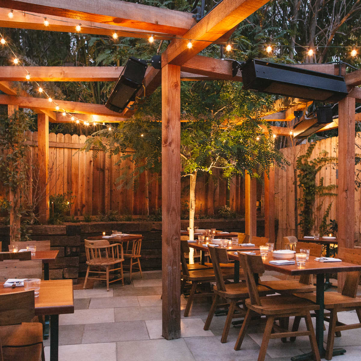 An outdoor restaurant patio with wooden tables and chairs arranged under string lights and surrounded by greenery.