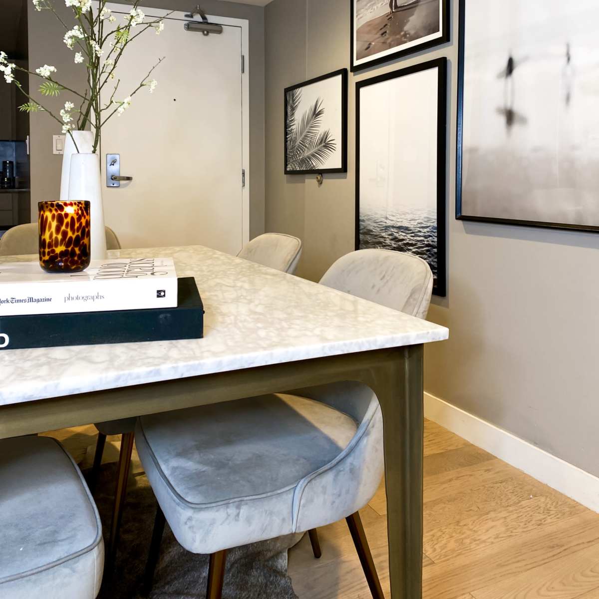 A dining area features a marble-top table surrounded by upholstered chairs, with art framed on the walls and a vase of branches as a centerpiece.