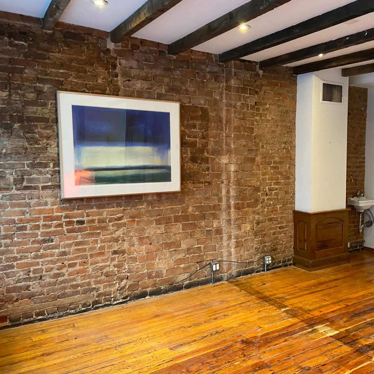 A room with exposed brick walls, wooden beams on the ceiling, and hardwood flooring, featuring a large framed artwork and a small bathroom fixture in one corner.