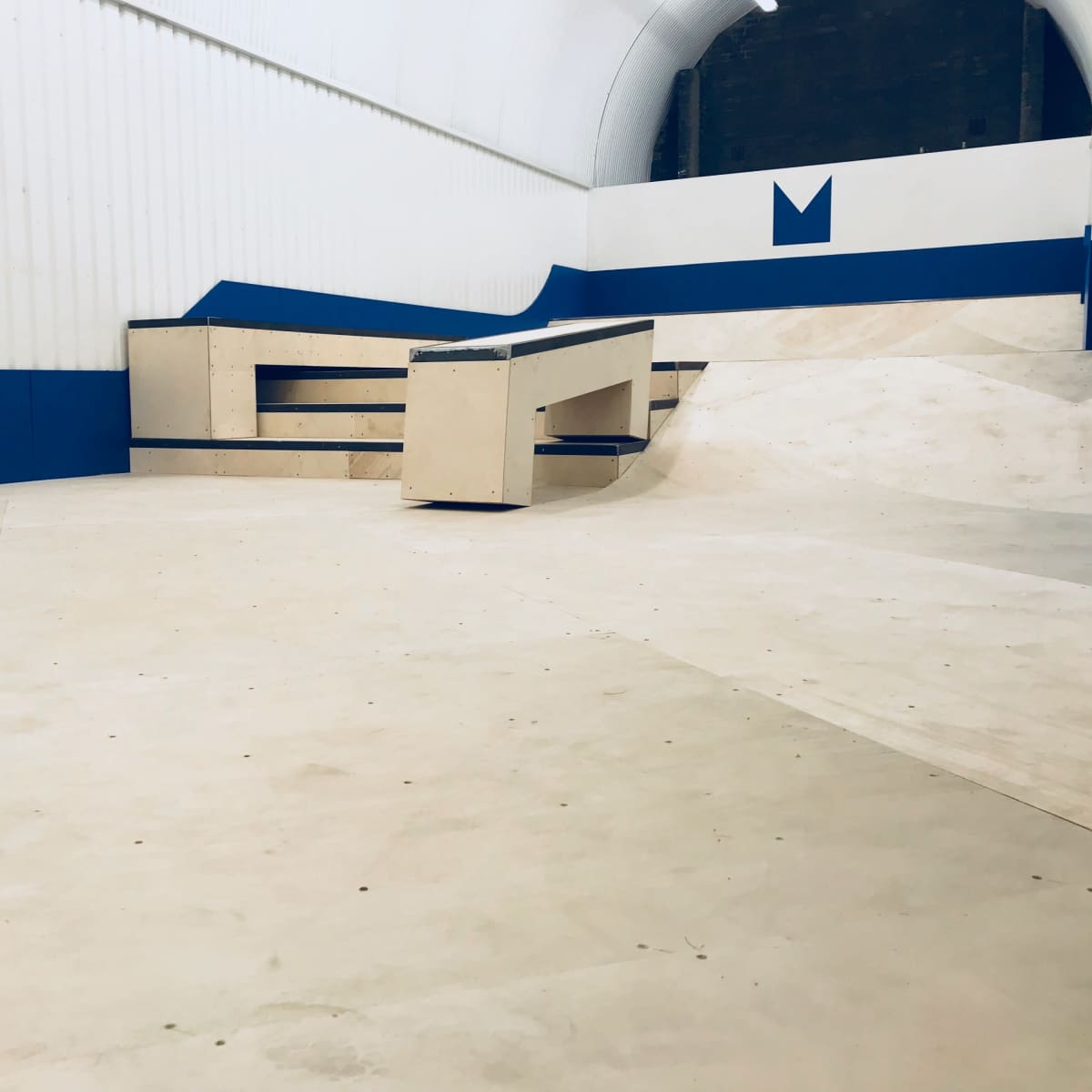 A skatepark with a smooth, light-colored floor, ramps, and a wooden obstacle surrounded by a blue and white wall.