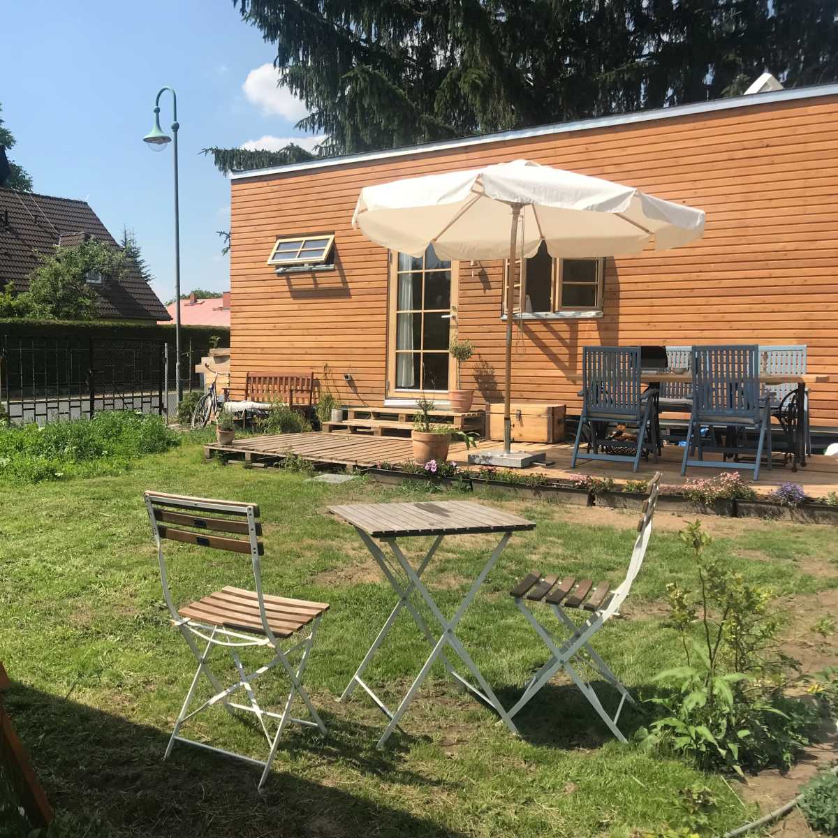 A wooden house is seen in a backyard with a swing set, patio furniture, and an umbrella on a sunny day.