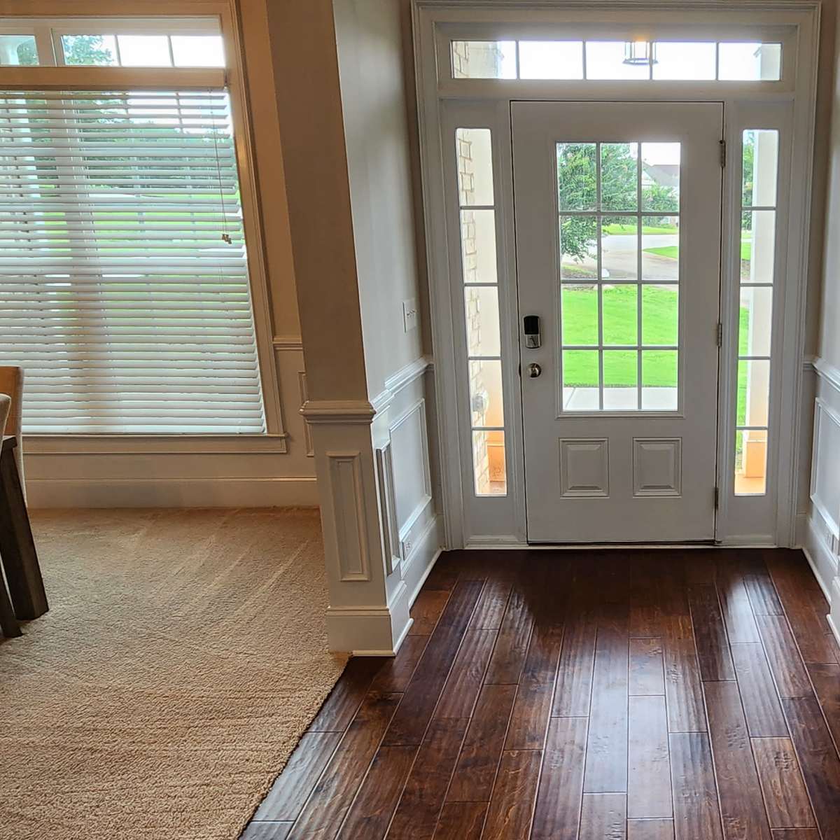 A hallway leading to a front door, with a large window on one side and a dining area with a table on the other, featuring wooden flooring and neutral-colored furnishings.