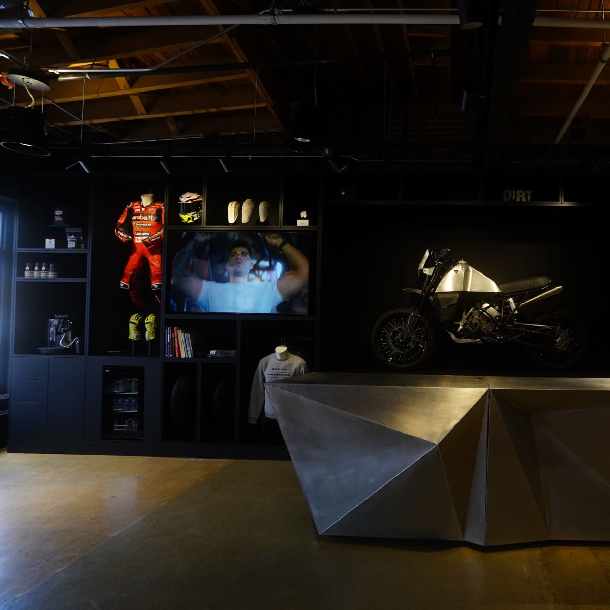 A modern retail space features a central metallic display with a motorcycle on top, surrounded by shelves displaying motorcycle apparel and accessories.