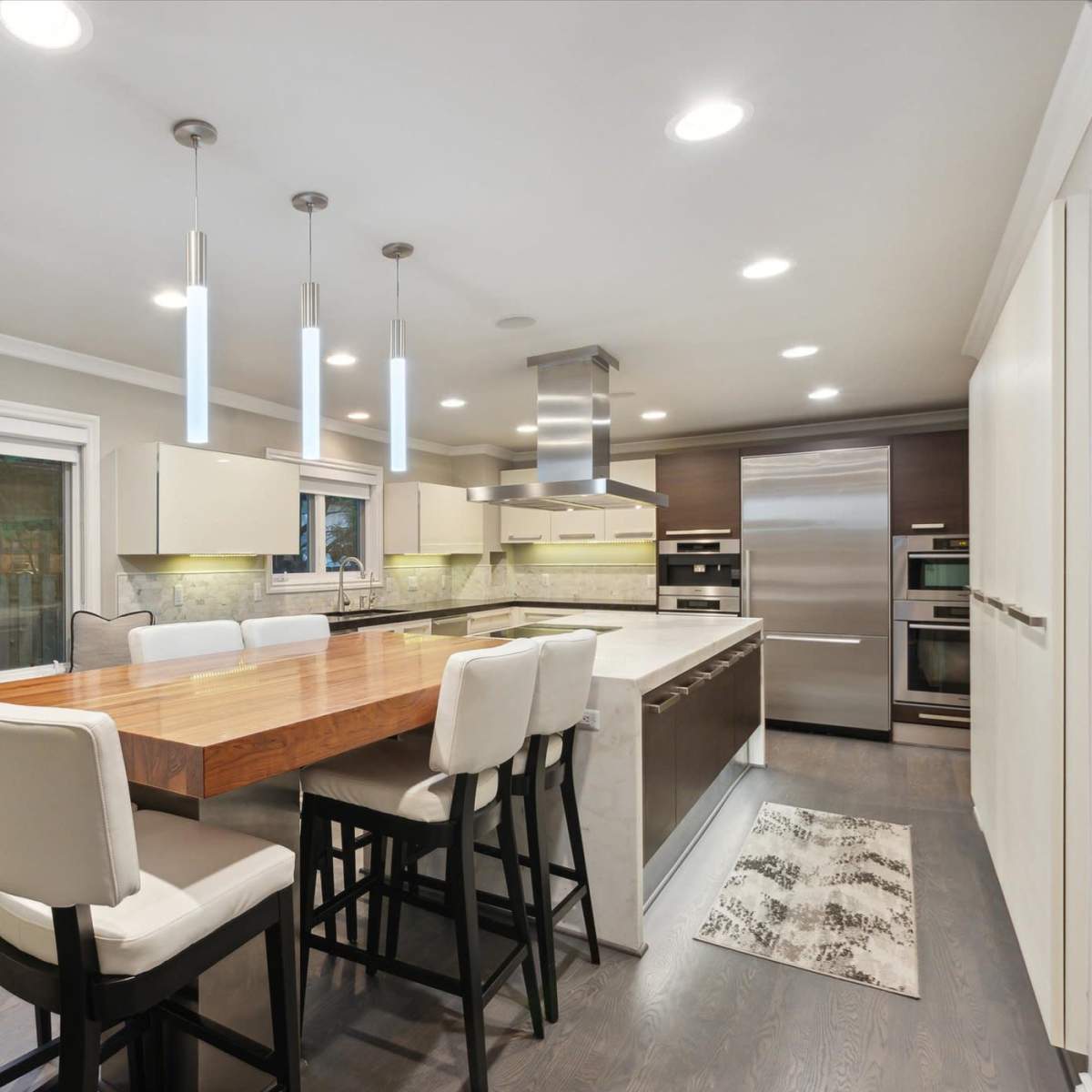 A modern kitchen features a large wood dining table with high-backed chairs, stainless steel appliances, and ample natural light from a side window.